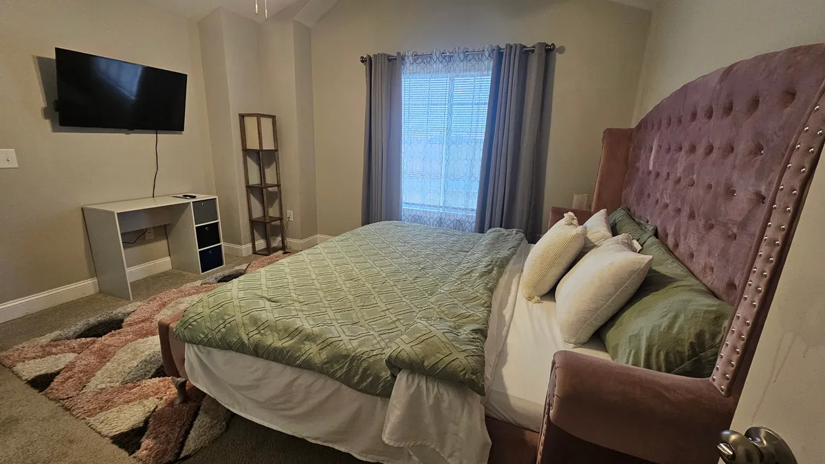 This interior bedroom scene centers on a large pink tufted headboard, inviting bed with green and white bedding, and a window dressed in sheer curtains. The overall atmosphere is intimate and tranquil, enhanced by natural light and a cozy setup ideal for relaxation and privacy.