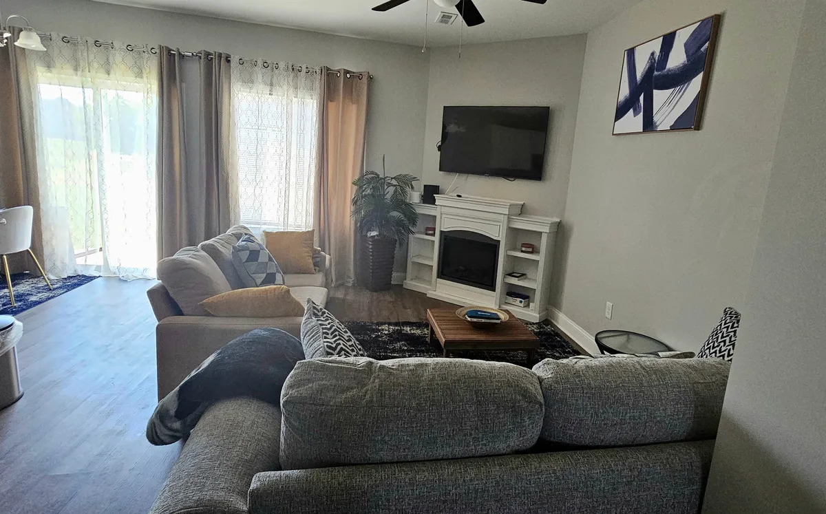 A bright and welcoming living room featuring comfortable sofas arranged around a coffee table, a large television as a focal point, and expansive windows that let in abundant natural light. The space is decorated with subtle, modern decor pieces and designed to foster both relaxation and connection.