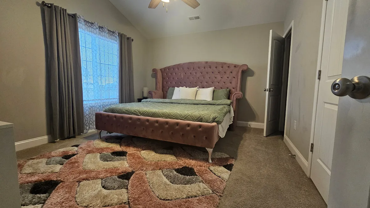 An inviting bedroom with a large pink tufted bed, a textured area rug, and a window letting in natural daylight. The setting exudes comfort and warmth, making it an appealing private space for rest and relaxation.