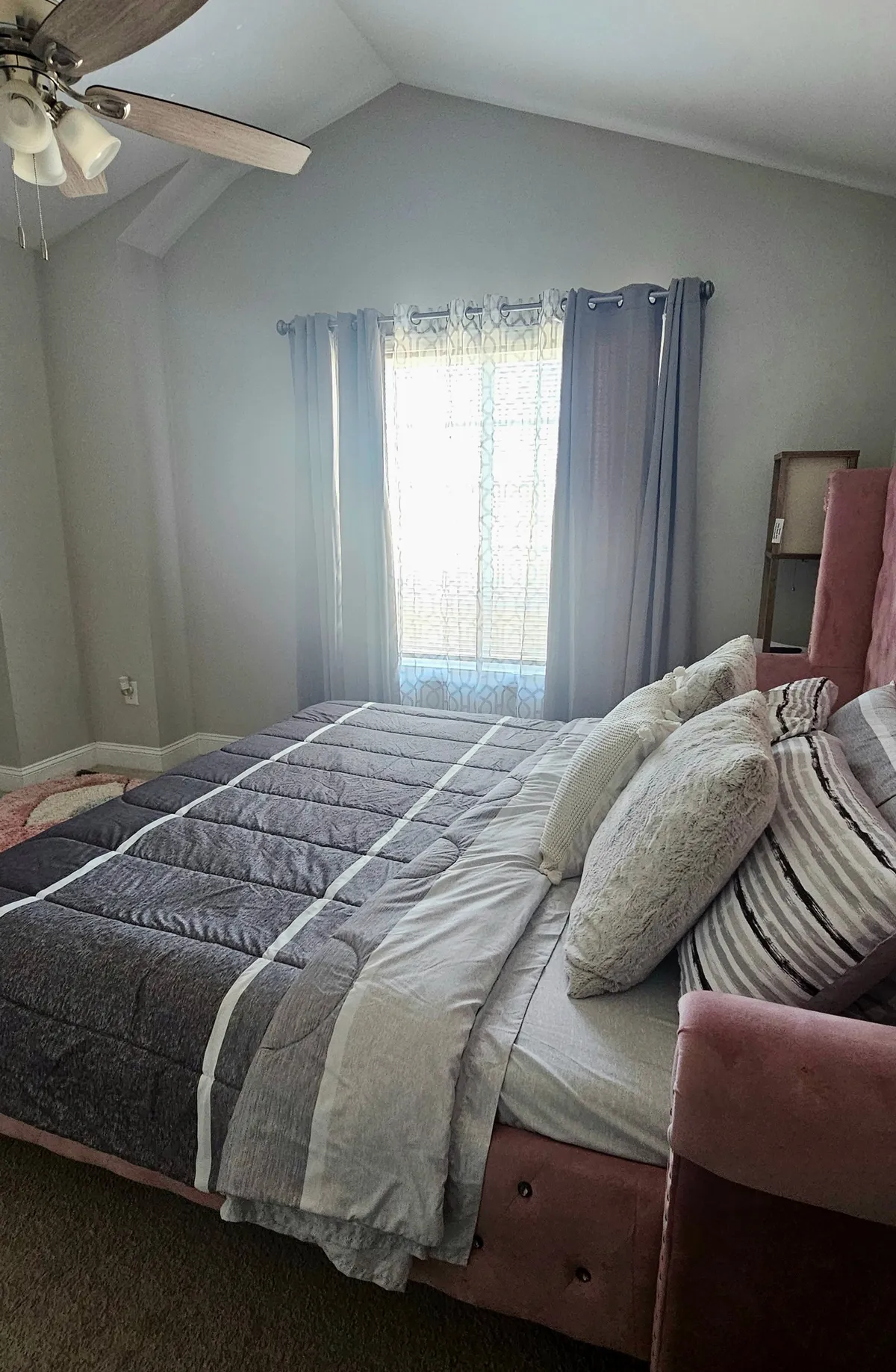 A calm, peaceful bedroom featuring a bed with decorative pillows, a window with sheer curtains allowing in gentle sunlight, and a ceiling fan. The room is softly lit, creating an inviting and restful atmosphere ideal for unwinding after a busy day.