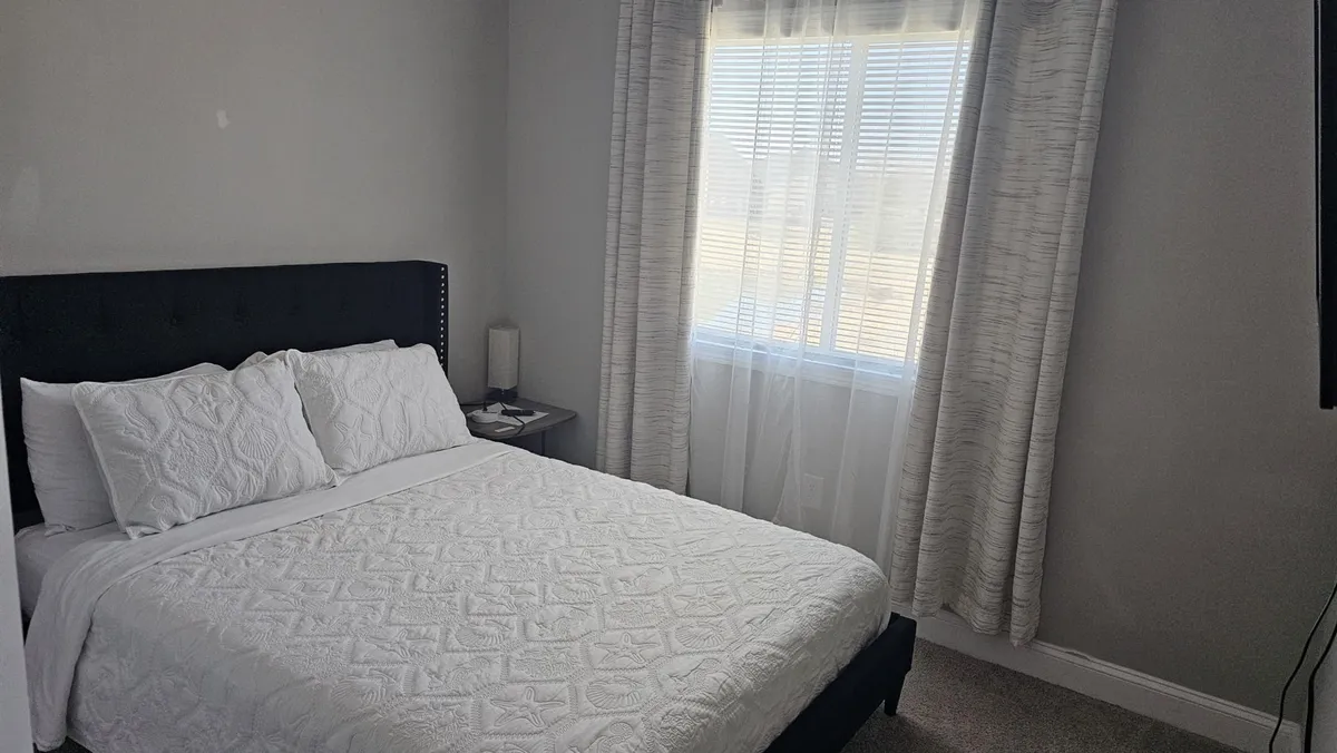 A tranquil bedroom with a neatly made bed, soft sunlight streaming in through sheer curtains, a cozy nightstand with a lamp, and a window-side reading nook. The scene exudes peacefulness, offering a glimpse of the home's relaxing interior retreat.